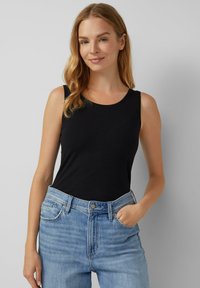 Mujer con pelo rubio, vistiendo una blusa negra sin mangas y jeans de color azul claro, de pie con una mano en el bolsillo, contra un fondo neutro.