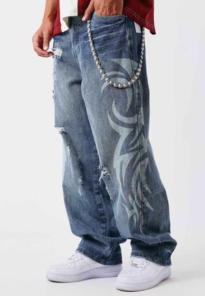 Wijde distressed jeans met een groot tribaal patroon op het been en een zilveren kettingaccessoire, gedragen met witte sneakers.