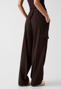 Bruine broek met wijde pijpen, zijzakken, een gladde textuur, een elastische tailleband en subtiele plooien aan de achterkant. Schoeisel is beige.