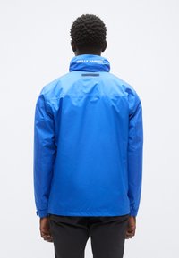 Μπλε αδιάβροχο μπουφάν με ψηλό γιακά, ελαστικά μανίκια και αεριζόμενη πλάτη. Διαθέτει λευκό λογότυπο στο λαιμό. Λείο, γυαλιστερό υφή.