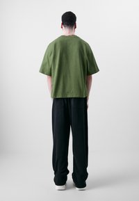Grønn bomulls oversized T-skjorte med korte ermer, baksiden har to knappdetaljer nær kanten, kombinert med svarte vide bukser.
