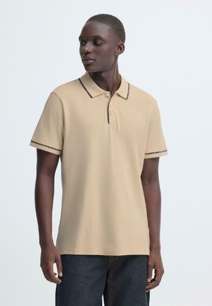 Homme portant un polo beige à manches courtes avec un liseré noir sur le col et les manches, debout devant un fond clair uni.
