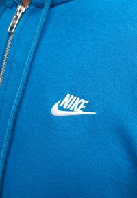 Sudadera azul hecha de tela suave, con un logotipo bordado de Nike en blanco en el lado izquierdo del pecho y un cierre plateado en la parte delantera.