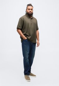 Άνδρας που στέκεται με το ένα χέρι στην τσέπη φορώντας χακί πόλο πουκάμισο, μπλε τζιν και καφέ αθλητικά παπούτσια μπροστά σε απλή ανοιχτόχρωμη φόντο.