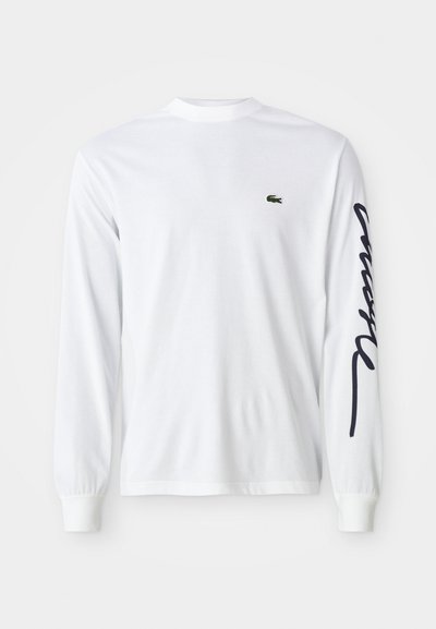 Chemise blanche à manches longues en coton, avec un logo brodé vert sur la poitrine et un texte scripté bleu foncé le long de la manche gauche.