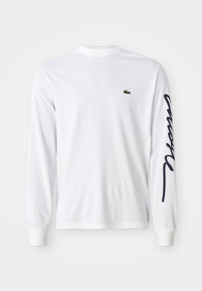 Chemise blanche à manches longues en coton, avec un logo brodé vert sur la poitrine et un texte scripté bleu foncé le long de la manche gauche.