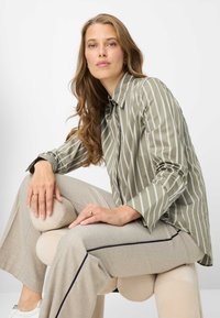 Olijfgroene gestreepte blouse met knopen en lange mouwen, gecombineerd met lichtbeige broek met wijde pijpen. Een gouden ring siert één hand.