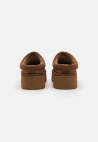 Brown fabric slip-on shoes with a round toe and a low, chunky heel. The texture appears soft with stitched detailing around the collar.