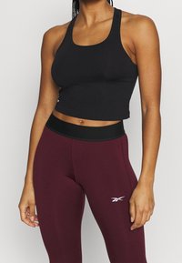 Žena v černém zkráceném topu a vysokých vínově červených legínách s černým páskem a bílým logem na stehně.