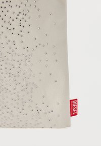 Μπεζ τσάντα tote με υφή διακοσμημένη με διάσπαρτα ασημ ένια τρουκς, με μια κόκκινη ετικέτα με το λογότυπο "DIESEL" στην άκρη.