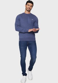 Marineblauwe sweatshirt met een ronde hals en lange mouwen, gecombineerd met blauwe slim-fit jeans en witte sneakers. Subtiele logodetail op de borst.