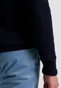 Marineblaue Strickjacke mit gerippten Bündchen und Saum, aus strukturiertem Stoff. Getragen mit hellblauen Cordhosen, Nahaufnahme des Ärmels.