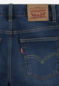 Jean en denim bleu avec une étiquette en cuir à la taille, des coutures jaunes et un design de poche arrière signature.