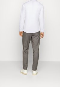 Camicia bianca a maniche lunghe con orlo arrotondato, abbinata a pantaloni grigi e sneaker bianche, vista da dietro. Design semplice e pulito.