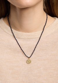 Perlekæde med sorte glasperler og et rundt guldfarvet vedhæng med indviklede mønstre og klare detaljer, båret over en beige sweater.