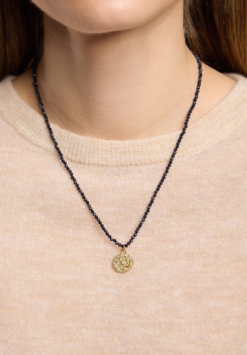 Perlekæde med sorte glasperler og et rundt guldfarvet vedhæng med indviklede mønstre og klare detaljer, båret over en beige sweater.