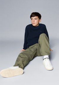 Granatowy sweter z dzianiny, oliwkowe dopasowane spodnie i białe sneakersy z zielonymi akcentami. Model siedzi na szarym tle.