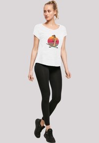 Hvit t-skjorte med grafisk trykk av en karakter mot en fargerik solnedgang, kombinert med svarte leggings og svarte treningssko.