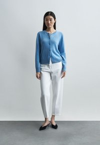 Γυναίκα που φοράει μπλε ζακέτα με κουμπιά, λευκό παντελόνι ψηλής μέσης και μαύρα μυτερά παπούτσια, στέκεται μπροστά σε απλό φωτεινό τοίχο.