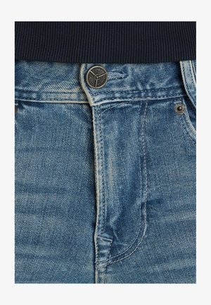 Jean en denim présentant un lavage bleu clair, des coutures blanches contrastantes et une fermeture à bouton en métal à la taille. Gros plan sur la texture du tissu.