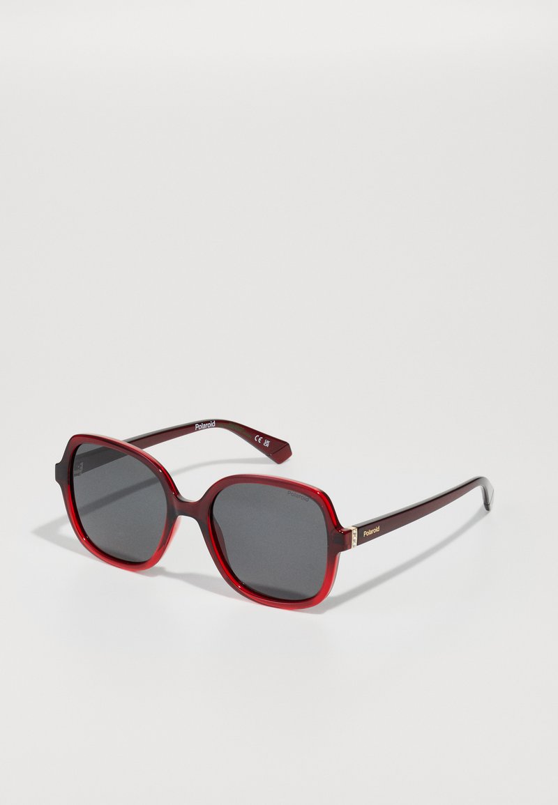 Óculos de sol com armações de plástico vermelhas, em formato quadrado, lentes cinza polarizadas e detalhe em dourado no braço.