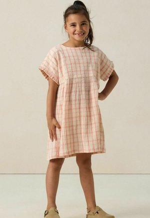 Fille souriante se tient les mains sur les hanches, portant une robe à carreaux rose et blanche et des chaussures à enfiler beiges sur un fond uni.