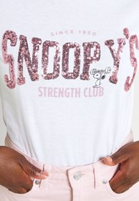 Valkoinen t-paita, jossa on vaaleanpunaisista niiteistä tehty teksti "SNOOPY'S STRENGTH CLUB" ja pieni sarjakuvahahmo, joka nostaa painoa.