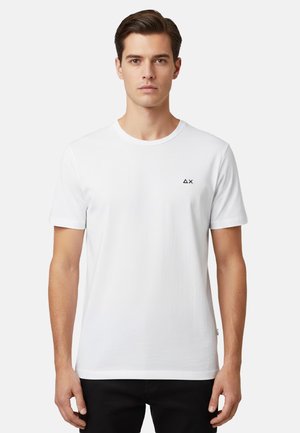 Giovane uomo che indossa una maglietta bianca a maniche corte con un piccolo logo a triangolo nero e una X sul petto sinistro, in piedi davanti a uno sfondo bianco.