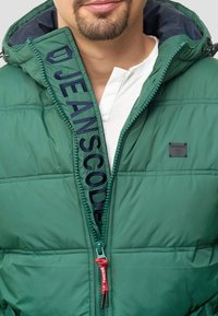 Giacca a vento verde con chiusura a zip e hardware rosso, grande ricamo sul colletto e una piccola etichetta logo sul petto.