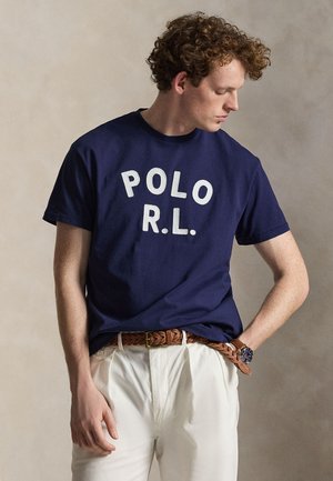 Junger Mann mit lockigem Haar, der ein dunkelblaues T-Shirt mit der Aufschrift „POLO R.L.“, weiße Hose, einen braunen geflochtenen Gürtel und eine Armbanduhr trägt.