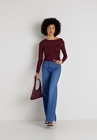 Burgundijas krāsas ilgu piedurkņu tops ar drapēta dizaina elementiem, apvienots ar gaiši ziliem, brīvi piegrieztiem džinsiem. Tur rokā mazu bordo krāsas rokassomu.