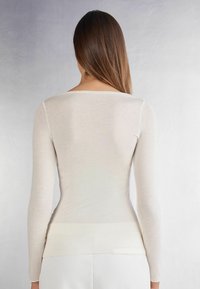 Cremfarbenes Langarmoberteil mit rundem Halsausschnitt, aus glattem Stoff gefertigt; hat eine figurbetonte Silhouette und einen etwas längeren Rückenabschluss.