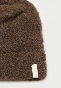 Béret tricoté marron avec une surface texturée et duveteuse. Il dispose d'une petite étiquette blanche sur le bord, affichant un design simple et fonctionnel.