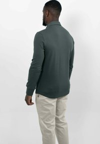 Donkergroene polo met lange mouwen en kraag, gecombineerd met lichtbeige broeken. Het shirt heeft een gebogen zoom en aansluitende manchetten.