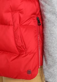 Rode puffer-vest met een gequilte textuur, voorzien van een ritszak en een rond logo. Het wordt gedragen over een lichtgrijze trui.