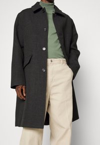 Man in donkere antraciet knoopjas over groen coltrui en beige wijde broek, staand met één hand in zijn zak.