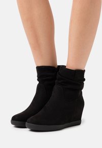 Bottes de cheville en suède noir à talon compensé, portées sur des jambes nues, avec un dessus légèrement plissé et un design à bout rond.
