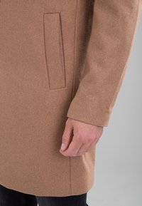 Bras et main droits d'une personne portant un manteau en laine couleur camel avec une poche fendue verticale sur la hanche, sur fond gris neutre.