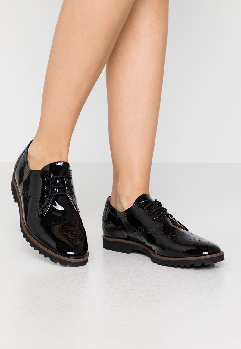 Femme portant des chaussures brogue en cuir noir brillant avec des perforations décoratives et des lacets noirs, debout sur un sol gris clair.