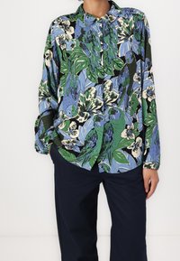 Bloemige blouse met lange mouwen, gekenmerkt door een levendig groen en blauw papegaaienpatroon op een zwarte achtergrond; zachte stof met een knopenkraag.