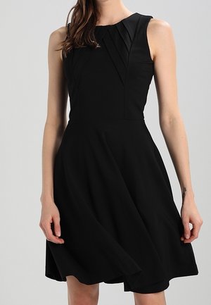 Femme portant une robe noire sans manches avec des détails plissés sur la poitrine et une jupe évasée sur un fond uni.