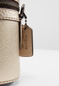 Złota torebka skórzana z fakturą, z ciemnym zamkiem błyskawicznym, z brązową metką z wygrawerowanym napisem "COACH", przymocowaną metalowym łańcuszkiem.