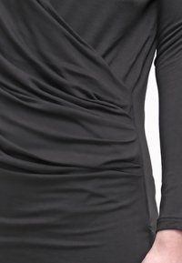 Czarna sukienka jerseyowa o dopasowanym kroju, z drapowaną tkaniną i delikatnym marszczeniem wzdłuż sylwetki. Długie rękawy z gładką teksturą.
