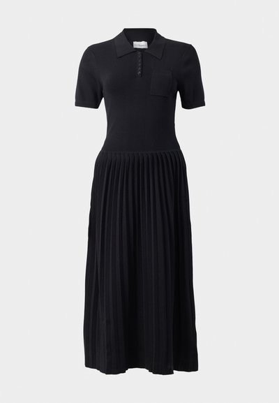 Robe polo noire à manches courtes, avec patte de boutonnage et poche poitrine. La jupe présente un design plissé, créant un aspect texturé.