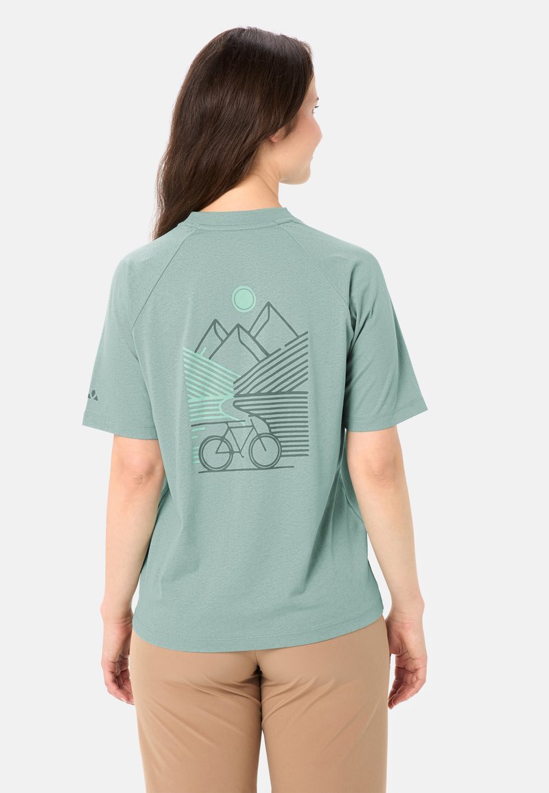 Vrouw met lang bruin haar draagt een lichtgroen T-shirt met een grafiek van een berg, zon en fiets op de achterkant, gecombineerd met beige broek.