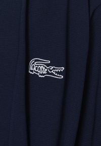 Tessuto strutturato blu navy con logo coccodrillo ricamato in bianco di Lacoste sul lato sinistro. Il design è minimalista e pulito.