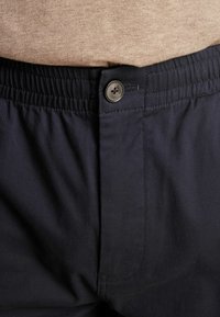 Gros plan sur un pantalon bleu marine avec taille élastique et fermeture à bouton, porté avec un haut beige rentré à l'intérieur.