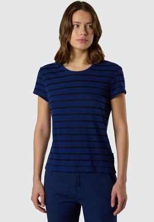 Femme aux cheveux bruns mi-longs portant une chemise bleu marine à manches courtes avec des rayures horizontales noires et un pantalon bleu marine, debout immobile.