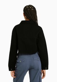 Cropad svart jacka med krage, har lång ärmar och knappmanschetter. Bärs med blå cargo-byxor som visar baksidan och fickor.