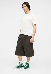 Νέος άνδρας που φοράει λευκό μπλουζάκι, σκούρα σορτς με φαρδιά πόδια, λευκές κάλτσες και πράσινα παπούτσια υψηλού πάτου, στέκεται σε ουδέτερη στάση.